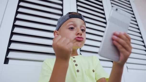 Retrato de un niño divertido mira en el teléfono, muestra un puño, infla sus labios, se enoja. Red de Internet, comunicación en línea, red social. Tecnología infantil y moderna — Vídeos de Stock
