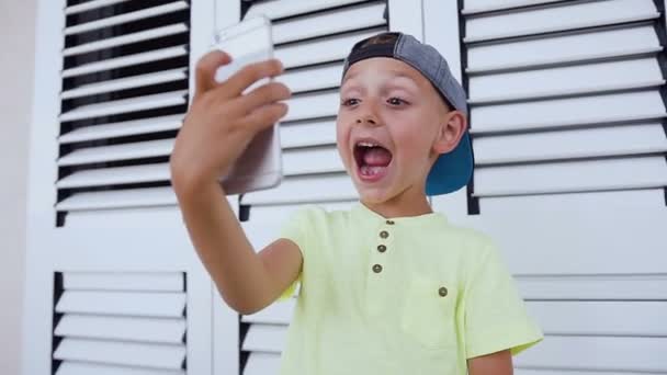 Divertido chico lindo con la boca abierta haciendo fotos selfies en su teléfono inteligente en casa, aislado sobre fondo blanco. Niño usando el teléfono móvil mientras juega juegos — Vídeo de stock