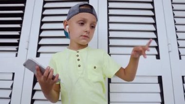 Gri smartphone kulaklıklardan müzik dinleyen şirin bir çocuk t-shirt ve şapka ve beyaz arka planda kapalı dans Close-up. Müzik dinlerken çekici bebek dans