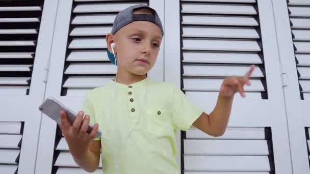 Primer plano de un lindo chico en camiseta y gorra que escucha música en auriculares desde el teléfono inteligente gris y bailando en el fondo blanco en interiores. Atractiva danza del bebé mientras escucha música — Vídeo de stock