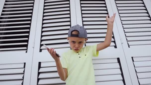 Een schattige kleine jongen te kijken naar de camera presteert geïmproviseerde dansen en genieten van de muziek die hij naar de koptelefoon van de telefoon luistert. Kind in t-shirt en cap is luisteren muziek vanaf een — Stockvideo