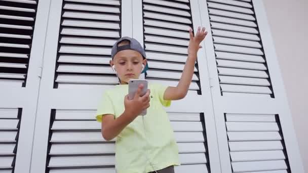 Retrato de um menino hipster em t-shirt e boné está ouvindo música favorita no smartphone com fones de ouvido dançando balançando a cabeça sobre fundo branco. Conceito de emoções — Vídeo de Stock