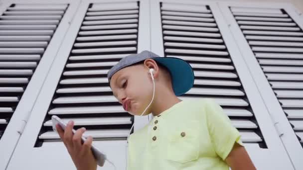 Un niño escucha música con auriculares desde un teléfono inteligente en el fondo blanco. Él cierra sus ojos y se sumerge en el mundo de la música bailando a un ritmo. Concepto de: tecnología y música, sueños y — Vídeo de stock