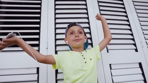 Niño divertido con auriculares está bailando y escuchando la música de un teléfono inteligente aislado sobre un fondo blanco — Vídeos de Stock