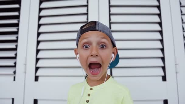 Retrato de menino hipster em boné com fones de ouvido que está ouvindo música favorita no smartphone com uma boca aberta e olhando para a câmera. Close-up, o garoto engraçado olha para a câmera — Vídeo de Stock