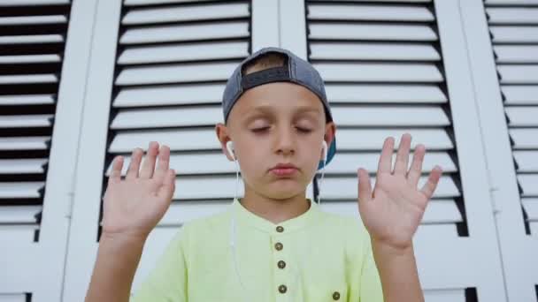 Ein lustiger kleiner Junge mit Kopfhörern, der Musik genießt, tanzt und gestikuliert isoliert vor weißem Hintergrund. Kind in Mütze und T-Shirt hört guten Song von seinem Smartphone — Stockvideo