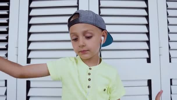 Retrato de un lindo niño con camiseta y gorra que escucha música favorita en el teléfono inteligente con auriculares, bailando agitando la cabeza sobre fondo blanco. Concepto de emociones . — Vídeos de Stock