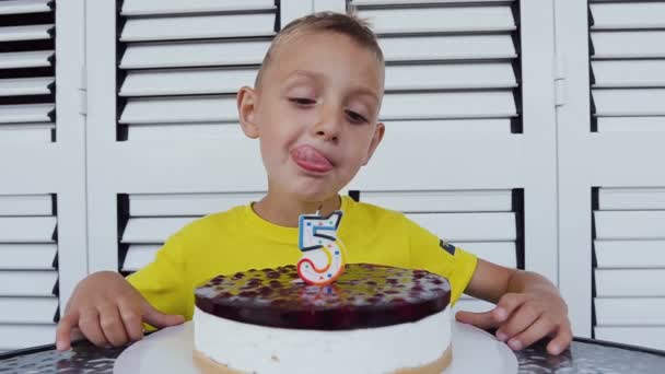 O menino bonito é celebração em seu quinto aniversário sentado na mesa com um delicioso bolo, isolado em um fundo branco. Retrato de um menino pequeno em seu aniversário — Vídeo de Stock