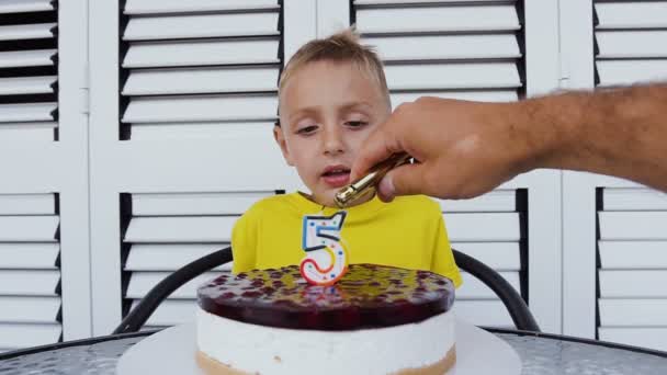 Niño sentado en la mesa y soplando velas en el pastel de cumpleaños. Lindo niño feliz celebrando su quinto cumpleaños, delante de él delicioso pastel con vela en forma de 5 — Vídeos de Stock