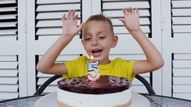 Bellissimo ragazzo di cinque anni in t-shirt, che festeggia il suo compleanno, soffia candele sulla torta al formaggio fatta in casa, coperta su sfondo bianco. Festa di compleanno per bambino — Video Stock