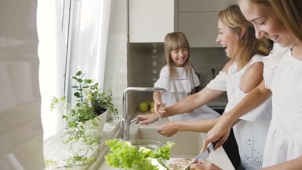 Junge kaukasische Mutter mit ihrer Tochter hat Spaß, spritzt sich gegenseitig Wasser in die Küche und wäscht das Gemüse für den fertig zubereiteten Salat zum Mittagessen — Stockvideo