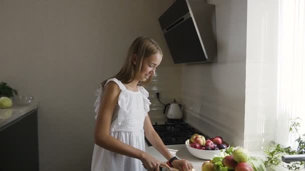 Menina adolescente atraente em vestido branco está preparando salada de frutas na cozinha. Menina consciente saudável está cortando frutas para uma refeição vegetariana em casa na cozinha — Vídeo de Stock