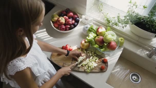 Vista superior. Chica rubia atractiva en vestido blanco está preparando ensalada de frutas en la cocina. La chica corta unas manzanas en su cocina en casa. Alimentación saludable — Vídeos de Stock