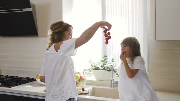 Bonito mãe e filha em roupas brancas fazendo salada com tomates na cozinha. Conceito alimentar saudável e vegan. Pequena menina atraente com cabelo longo come saborosos tomates maduros na cozinha — Vídeo de Stock