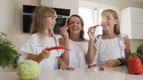 Nahaufnahme. attraktive Mutter mit ihren beiden Töchtern schneidet sich einen Schnurrbart aus rotem Paprika und hat Spaß dabei, zu Hause in der Küche lächelnd vor der Kamera zu posieren — Stockvideo