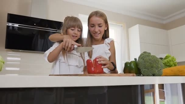 Dos chicas adolescentes en vestido blanco cocinan en la cocina: Las chicas cortan pimientos rojos para hacer una ensalada. La hermana mayor enseña a la hermana menor a cortar pimentón. Las chicas preparan ensalada de verduras en casa en el — Vídeos de Stock