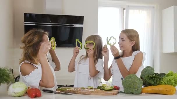 Jovem mãe com duas filhas bonitas está em conjunto preparando salada em casa cozinha theyre brincando com fatias de pimenta no conceito de nutrição vegetal saudável. Feliz bonito família brincando no — Vídeo de Stock