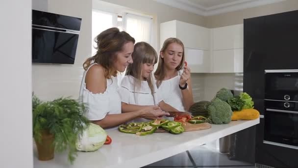 Mãe feliz e duas filhas estão cozinhando legumes para o jantar em casa cozinha. Comida, alimentação saudável, família — Vídeo de Stock