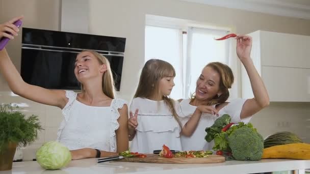 Ελκυστική χαμογελαστή γυναίκα με δύο κόρες κάνουν κάνοντας selfies, φωτογραφίες στο smartphone στην κουζίνα στο σπίτι. Τα κορίτσια φωτογραφίζονται με τα λαχανικά στο smartphone στην κουζίνα στο σπίτι — Αρχείο Βίντεο