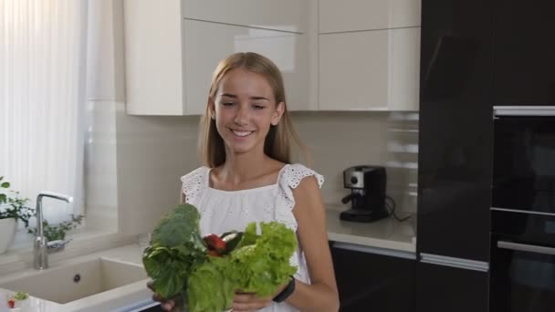 Привлекательная блондинка в белом платье несет стеклянную тарелку с овощами в руках на кухне. Мама и две дочери учатся вместе резать овощи на кухне, используя доску для рубки и — стоковое видео
