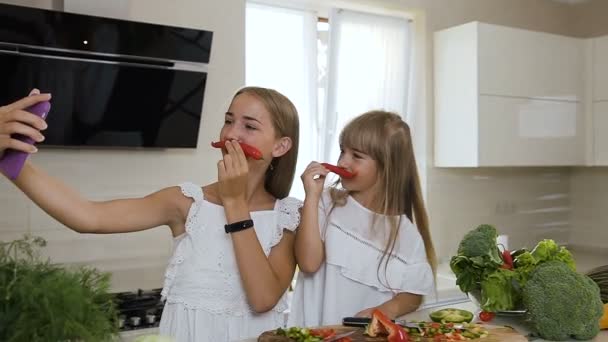 Две сестры с длинными волосами, одетые в белую одежду, показывают усы от красного перца чили и делают селфи фото на смартфоне дома на кухне. Технологии, люди, кулинария — стоковое видео
