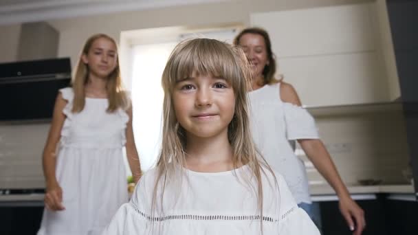 The charming girl with long hair in white dress stands and looks in the camera, mother and older sister fit hug and kisses her at home in the kitchen. Portrait of a lovely family — Stock Video