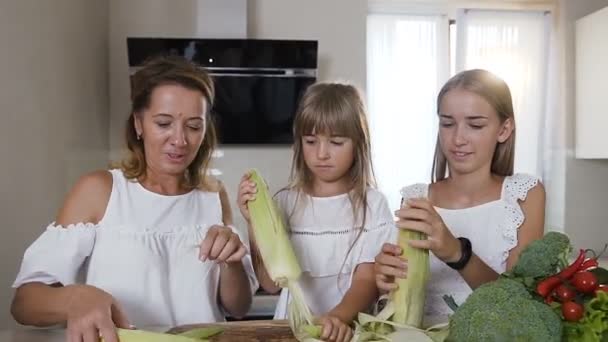 Крупним планом молода кавказька жінка з двома дочками в білому одязі прибирає, шльопає і готується готувати свіжу кукурудзу на кухні вдома — стокове відео