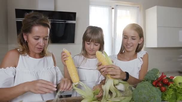 Atractiva mamá y sus hijas limpiando las espigas maduras de maíz de las hojas en la cocina del hogar. La familia feliz está limpiando el maíz de las hojas para cocinar. Alimento saludable — Vídeo de stock