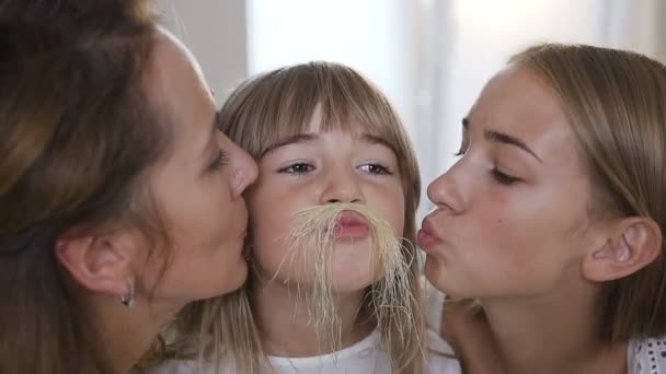 Närbild charmig liten flicka med långt hår gör en mustasch från majs hår och ser på kameran, Mamma och storasyster kyssa hennes kind hemma i köket. Porträtt av en härlig familj — Stockvideo