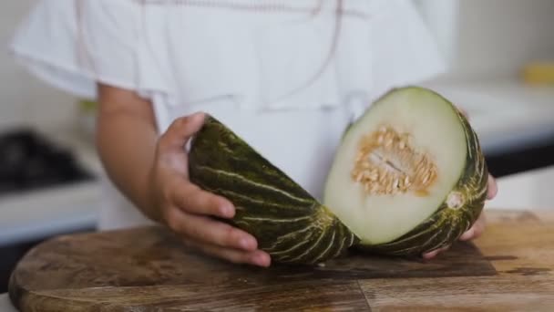 Cute dziewczynka w białej sukni trzyma w dłoniach, melon pokroić na pół w domowej kuchni. Zbliżenie: split zielony melon w połowie na płycie w rękach dziewczynki — Wideo stockowe