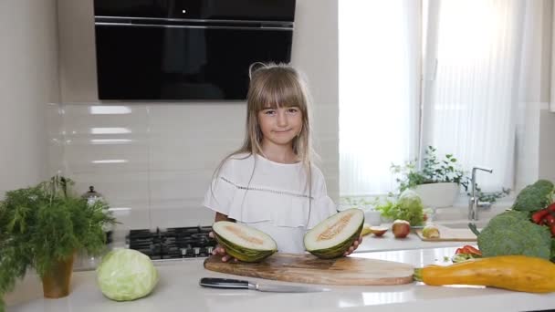 A menina bonito em vestido branco em uma tábua de madeira detém um melão verde cortado ao meio na cozinha da casa. Comida saudável, vegetal, criança — Vídeo de Stock