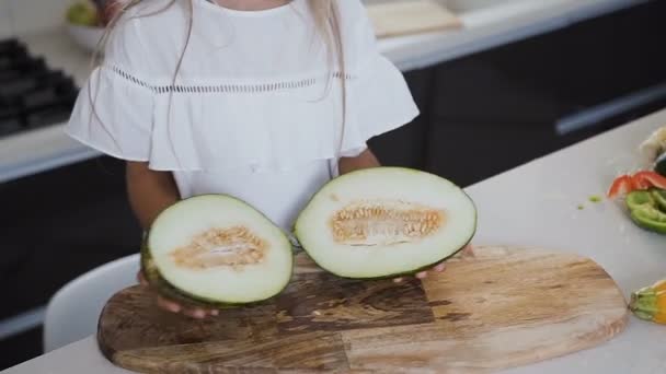 La niña en vestido blanco sostiene en sus manos un melón cortado por la mitad en la cocina del hogar. El corte de cerca del melón verde partido por la mitad en una tabla de madera en las manos de una niña — Vídeo de stock