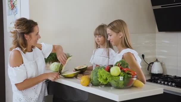 Madre e hijas cortando y comiendo melón verde en la superficie de trabajo de la cocina en casa. La mujer está cortando un melón y luego lo come con sus hijas — Vídeo de stock