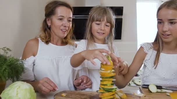 Carino bambina con la mamma e la sorella maggiore vestita con abiti bianchi divertirsi fa una piramide con zucchine al tavolo della cucina a casa in cucina. Famiglia felice, cibo sano, verdure — Video Stock