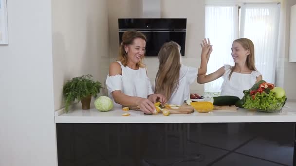 Zwei süße Schwestern und ihre schöne Mutter mit langen Haaren in weißen Kleidern geben sich gegenseitig die High Five, während sie zu Hause in der Küche kochen. kleines Mädchen und ihre Mutter mit ihrer älteren Schwester haben Spaß in — Stockvideo