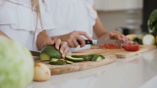 Duas adolescentes de roupas brancas estão cortando legumes em uma tábua de madeira atrás da mesa da cozinha na cozinha em casa. Corte de pimenta vermelha e abacate para cozinhar alimentos — Vídeo de Stock