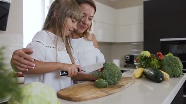 Femme attrayante souriante avec sa fille coupé moitié brocoli frais sur une planche à découper en bois. La petite fille aide sa mère à cuisiner la soupe. Mode de vie sain — Video