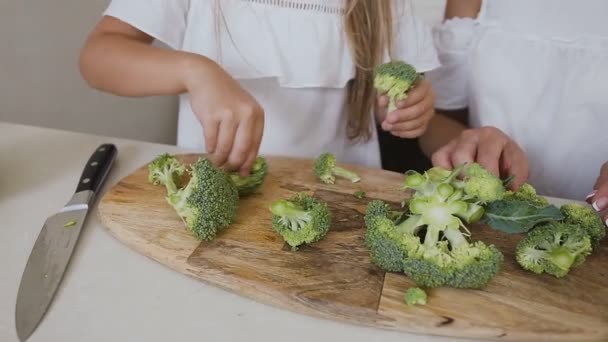Kleines Mädchen mit ihrer Mutter, die frischen Brokkoli auf einem Schnitzbrett schneidet. Nahaufnahme der Hände des Weibchens mit Brokkoli-Scheiben, die sich zu Hause in der Küche auf das Essen vorbereiten. Gesunder Lebensstil. Gemüse — Stockvideo