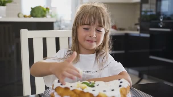 Retrato de uma menina adorável comendo bolo doce fresco em casa. Menina encantadora com cabelos longos em um vestido branco sentado na mesa da cozinha, comer é um delicioso bolo com creme e olhando para o — Vídeo de Stock