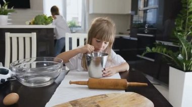 Biraz sevimli bebek kız beyaz elbiseli uzun saçlı mutfak masada oturuyor ve annesi mutfakta meşgul ise un karışık. Un bir kase ve beat yapmak meyilli içine karışık