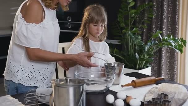 Atraente jovem mãe preparar tudo para cozinhar, derramando a farinha na tigela de vidro e sua filha ajudá-la. Mulher branca bonita com sua filha cozinhar na cozinha em casa — Vídeo de Stock