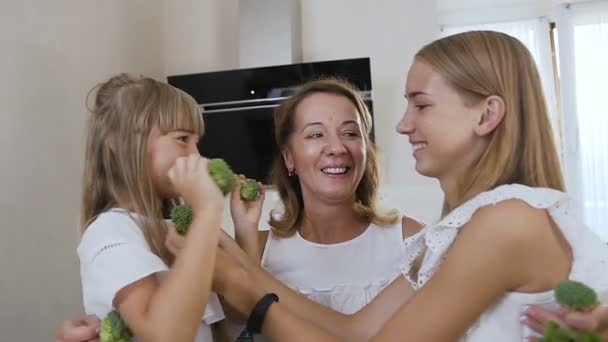 Close up do abraço apertado da bela mãe e suas duas filhas durante a culinária na cozinha em casa. Tiro de retrato. Família alegre brincando com legumes na cozinha, comida saudável — Vídeo de Stock