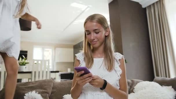 Beautiful caucasian student girl sits on bed using smartphone and smiling her little sister have fun jumps behind her with a soft cushion on the couch at home. Happy family. Concept of technology — Stock Video
