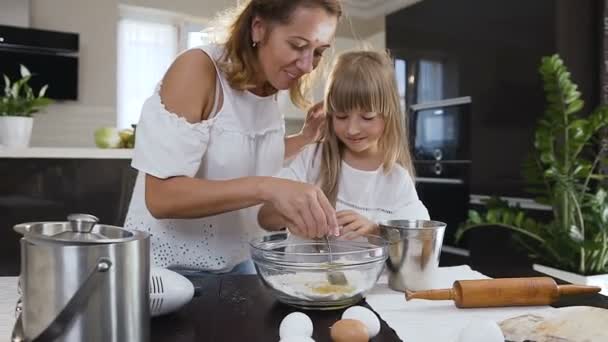 スプーンでケーキの材料を攪拌によって台所で彼女のお母さんを助ける少女。ボウルにクッキーの生地を攪拌しながら台所で母親を助けるかわいい女の子 — ストック動画