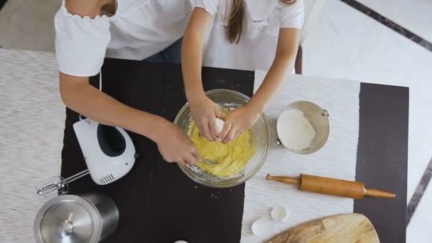 Вид сверху. Маленькая девочка помогает своей маме на кухне разбивать яйца в миску и смешивать тесто для печенья. Концепция семьи, еды, дома и людей — стоковое видео