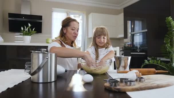 Поездка на камеру счастливых улыбок матери и дочери месить тесто вместе в большой стеклянной чаше на кухне. Счастливая семья на кухне. Мать и ребенок готовят тесто, пекут — стоковое видео
