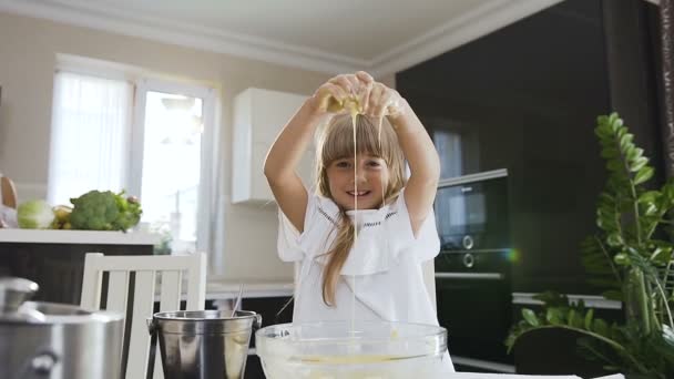 Linda niña con el pelo largo en vestido blanco amasando la masa en un tazón de cristal y mira a la cámara en la cocina moderna en casa. Las manos de los niños están sucias con masa. Chica hornear y mezclar masa de harina — Vídeo de stock