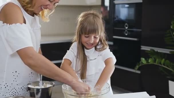 In Großaufnahme kneten die glücklich lächelnde Mutter und ihre kleine Tochter in der großen Glasschüssel in der Küche gemeinsam einen Teig. Kleines Mädchen hilft ihrer Mutter Teig zu kneten — Stockvideo