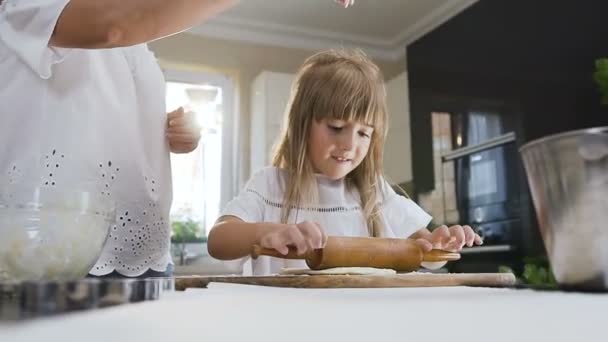 Good looking mother teaching her young little daughter to cook and kneading a dough for cookies on the table in the kitchen at home. Young woman and small girl preparing pasta or pizza together using — Stock Video