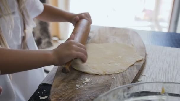 Chiudi u della graziosa bambina facendo un impasto e rotolandolo sul tavolo di legno ricoperto di farina con un mattarello in cucina a casa. All'interno. Ragazza rotolamento pasta cottura biscotti — Video Stock